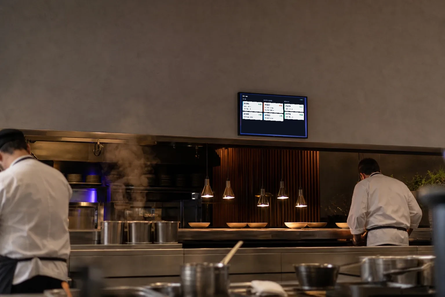 A kitchen display screen glowing above the pass in a busy professional restaurant kitchen.